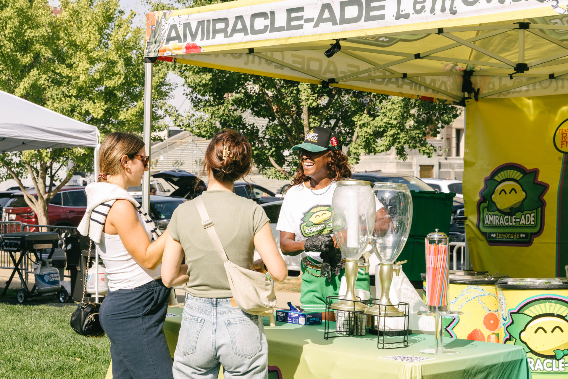 A lemonade vendor in a green cap and gloves smiling as she serves two customers at a bright outdoor market stand